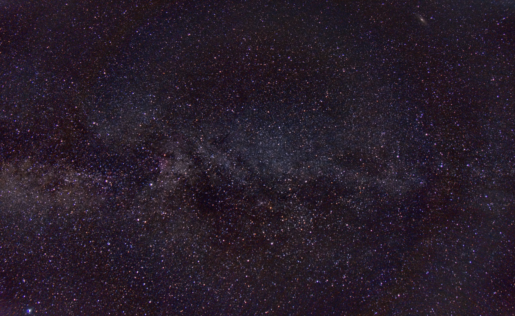 Deep stack of the Milky Way: a dense star field crossed by a pale band of starlight, with the Andromeda Galaxy as a small elongated smudge at upper right